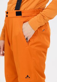 Les pantalons de ski orange présentent une texture lisse, une taille élastique avec des boutons et du Velcro, ainsi que des poches zippées. Un logo est présent sur le côté inférieur.