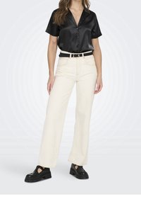 Vrouw in een zwart satijnen shirt met korte mouwen, hoog geplaatste off-white wijde broek met zwarte riem en zwarte chunky schoenen, staand tegen een neutrale achtergrond.