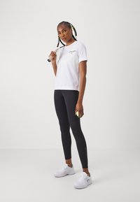 T-shirt branca de algodão com mangas curtas, combinada com leggings atléticas pretas e ténis brancos, segurando uma raquete de ténis e uma bola.