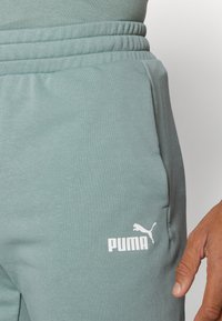 Šviesiai žali sportiniai džinsai, pagaminti iš minkštos medžiagos, turintys elastingą juosmenį ir balta Puma logotipą ant kairio šlaunies. Lygi tekstūra, kasdiens dizainas.