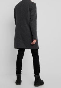 Cappotto di lana grigio, di lunghezza media, a taglio dritto, con un colletto alto e polsini abbottonati. Indossato con jeans neri e stivaletti neri.