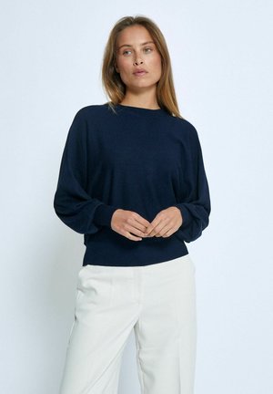Maglione pullover di un blu navy scuro con scollatura rotonda, maniche abbassate e polsini a costine. Tessuto liscio, vestibilità morbida, abbinato a pantaloni chiari.