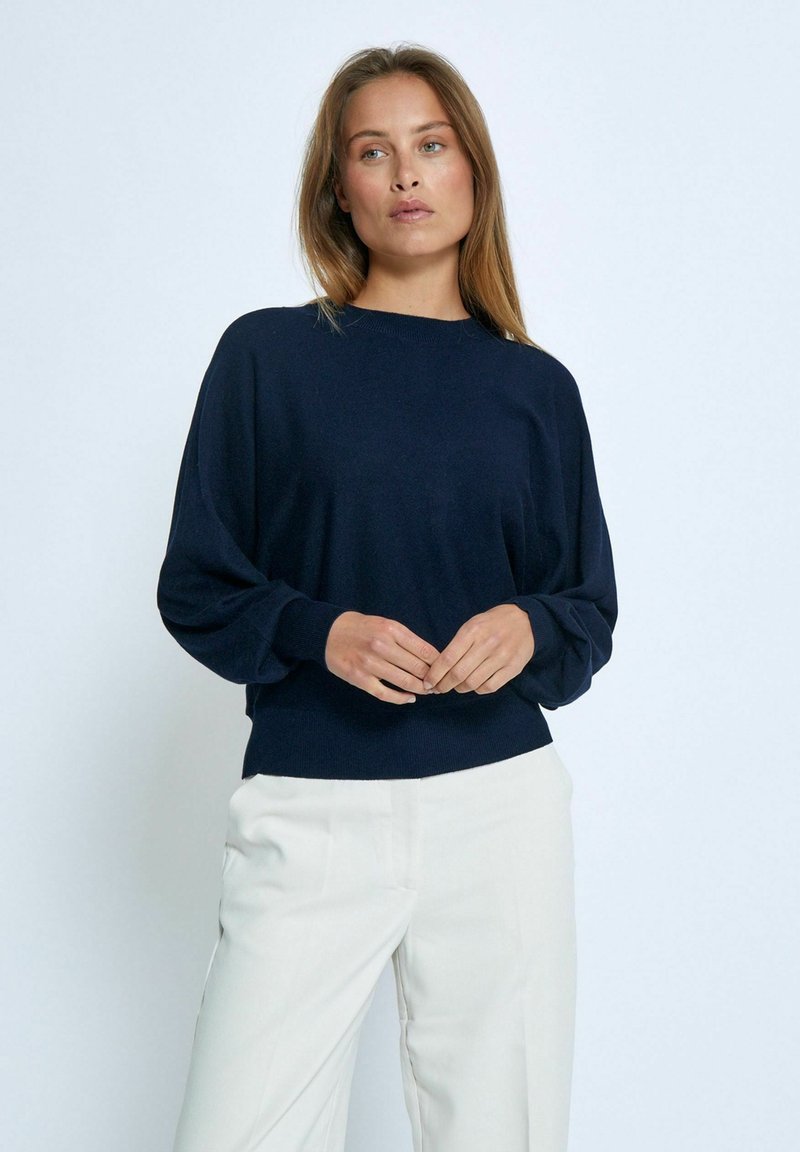 Dunkelblauer Pullover mit Rundhalsausschnitt, überschnittenen Schultern und gerippten Bündchen. Glatte Textur, lockere Passform, kombiniert mit hellen Hosen.