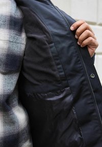 Schwarze Jacke mit Reißverschluss und kariertem Innenfutter. Verfügt über weiche Fleece- und glatte Stofftexturen. Entworfen mit lockerer Passform und Taschen.