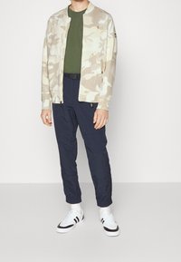 Giacca bomber da uomo con stampa camo beige, abbinata a una T-shirt verde oliva e pantaloni blu navy. Sneakers bianche con dettagli neri completano il look.