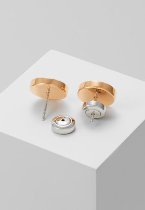 Deux boucles d'oreilles rondes de couleur or avec un poussoir argenté retiré et placé à côté sur une surface blanche.