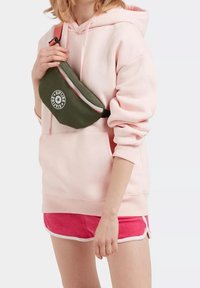 Sac banane kaki avec un logo rond, porté sur un hoodie rose clair et un short rose avec des bordures blanches. Le matériau semble lisse et durable.