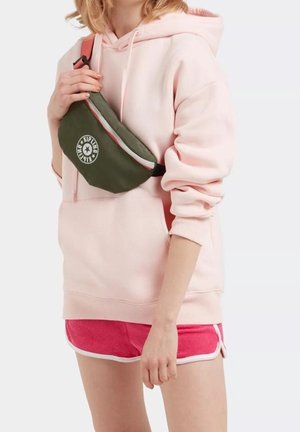 Sac banane kaki avec un logo rond, porté sur un hoodie rose clair et un short rose avec des bordures blanches. Le matériau semble lisse et durable.