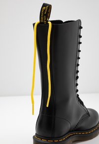 Botte en cuir noire à lacets jaunes et coutures jaunes, avec une languette noire et jaune avec texte en haut à l’arrière.
