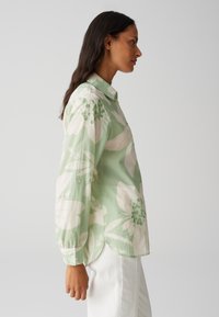 Blusa floreale verde chiaro con grandi fiori bianchi e dettagli verdi, stile con bottoni, vestibilità ampia, maniche lunghe e spacchi laterali.