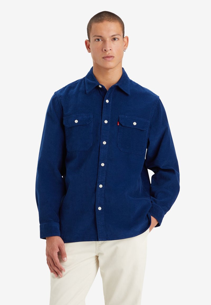 Camisa de pana azul marino con bolsillos en el pecho abotonados, mangas largas y botones blancos. Presenta un cuello y un tejido suave y texturizado.