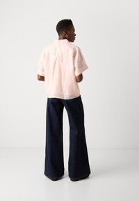 Camicia rosa chiaro a maniche corte con piega sul retro, abbinata a pantaloni in denim a gamba larga blu scuro. Design semplice con maniche arrotolate e senza motivi visibili.