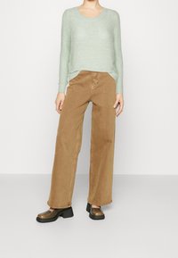 Osoba oblečená v světle zeleném pleteném svetr, širokých hnědých kalhotách a tmavě hnědých podpatcích, stojící proti obyčejné zdi.