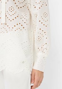 Weiße Bluse mit filigraner Lochstickerei, abgerundetem Saum und langen Ärmeln. Verfügt über Knopfdetails und gewellte Kanten. Leichter Stoff.