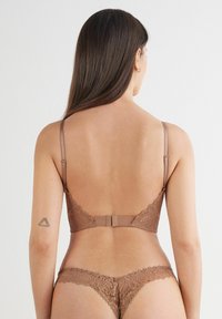 Brązowy koronkowy stanik z regulowanymi ramiączkami i zapięciem na haczyk. Koronkowe brzegi w kształcie falbany na miseczkach oraz pasujące koronkowe stringi, oba z misternymi wzorami.