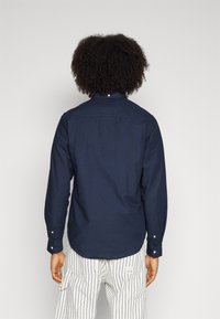 Navy blue long-sleeve shirt with buttoned collar and cuffs, featuring a smooth texture and a curved hem, worn over striped pants.