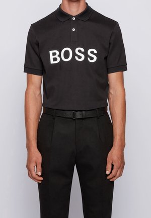 Sort polo skjorte lavet af bomuld med korte ærmer, med en fremtrædende hvid "BOSS" logo på brystet. Kombineret med sorte bukser.