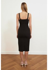 Robe midi noire ajustée avec larges bretelles, fermeture éclair au dos et texture lisse, associée à des sandales à talons hauts couleur nude sur un sol en bois.