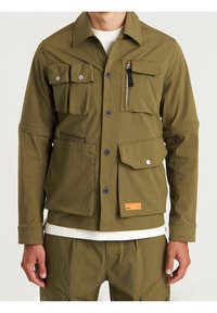 Mężczyzna w oliwkowym, roboczym płaszczu z wieloma kieszeniami z przodu, z guzikami, detalami na zamek oraz pasującymi spodniami.