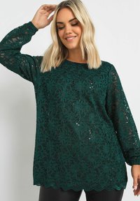 Långärmad topp i mörkgrön spets med blommönster och paljetter. Har en vågig nederkant och en lös passform. Slät textur, lätt design.