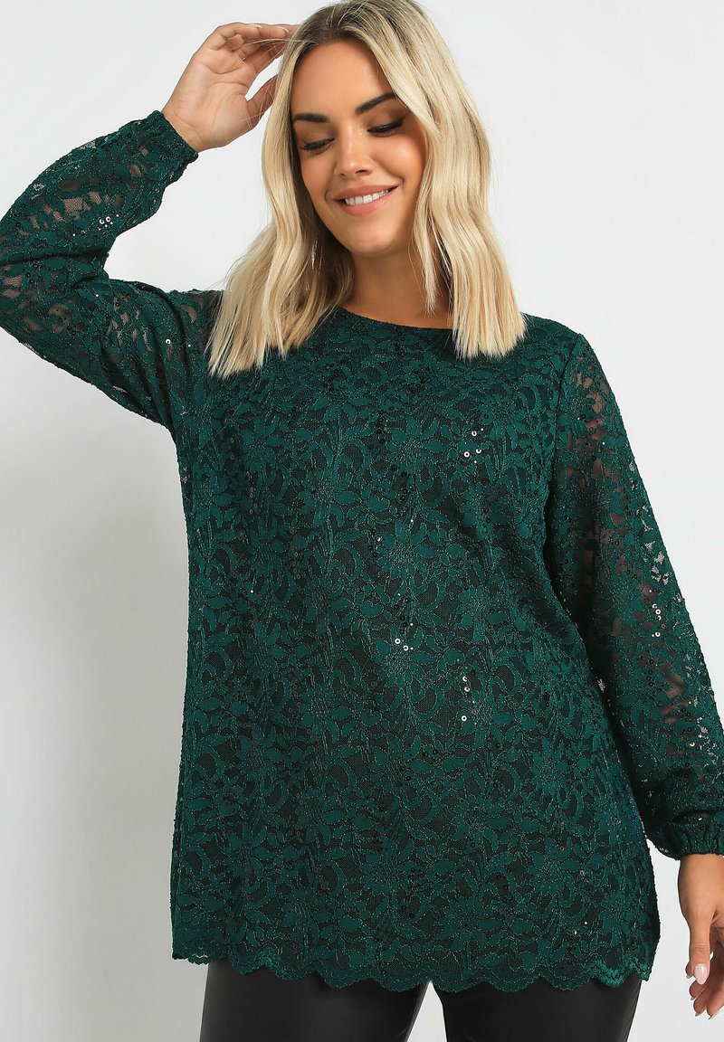 Långärmad topp i mörkgrön spets med blommönster och paljetter. Har en vågig nederkant och en lös passform. Slät textur, lätt design.