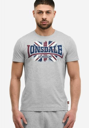 Uomo che indossa una maglietta grigio chiaro e pantaloncini; la maglietta mostra una Union Jack usurata con la scritta "Lonsdale London".
