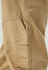 Beżowe spodnie cargo z boczną kieszenią, wykonane z materiału bawełnianego, z widocznym ściegem i gładką fakturą. Cechuje je luźny krój i funkcjonalny design.