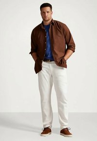 Camisa castanha com botões por cima de uma t-shirt azul, combinada com calças brancas e sapatos castanhos. A camisa tem um corte descontraído e mangas enroladas.