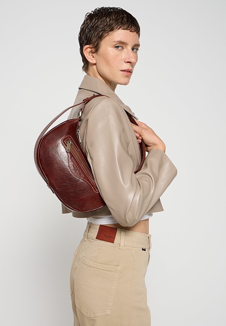 Brązowa skórzana torba na ramię o zaokrąglonym kształcie, wyposażona w kieszeń na zamek błyskawiczny i wykończenia w kontrastowym kolorze, noszona przez modelkę ubraną w beż.