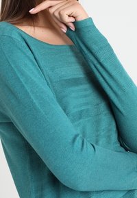 Teal knit sweater featuring a textured design and long sleeves, with a wide neckline and a relaxed fit.