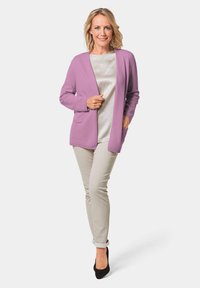 Lavendelfarbenes langes Strickcardigan mit offenem Vorderteil, zwei Vorderentaschen, getragen über einem hellen Satinoberteil und beigen Hosen, kombiniert mit schwarzen High Heels.