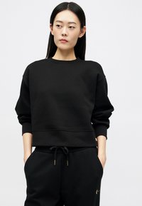 Νεαρή γυναίκα με μακριά ίσια μαλλιά φοράει μαύρη μπλούζα και μαύρο παντελόνι με κορδόνια, με τα χέρια στις τσέπες, κοιτάζοντας ελαφρώς στο πλάι.