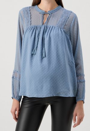 Kvinde iført en lyseblå gennemsigtig bluse med blonde detaljer og bindebånd ved halsudskæringen, kombineret med sorte bukser i kunstlæder.