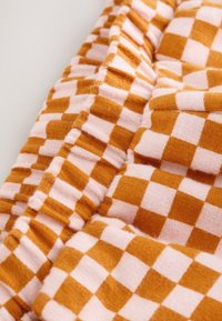 Tissu de coton présentant un motif à carreaux orange et rose clair. La texture est douce avec un détail froncé le long du bord.