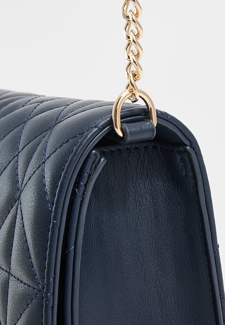 Bolso de mano de piel acolchada en azul marino con cadena dorada y anilla de sujeción, mostrado en primer plano en el lateral y el borde superior.