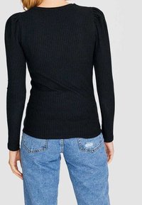 Kvinna som bär en åtsittande svart ribbad långärmad tröja och ljusblå högmidjade jeans, stående vänd bort.