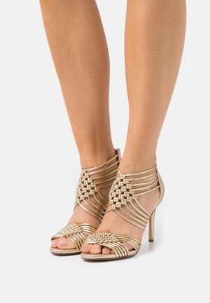 Gold strappy high-heeled sandals with beaded accents, featuring a crisscross design and open toe, set against a neutral background.