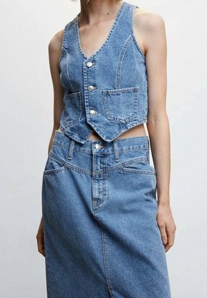 Femme portant un gilet bleu en denim sans manches boutonné et une jupe en denim taille haute assortie avec passants pour ceinture, sur fond uni.