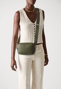 Olive green crossbody bag with a rectangular shape, gold hardware, and a textured strap. Worn over a cream knit top and light pants.