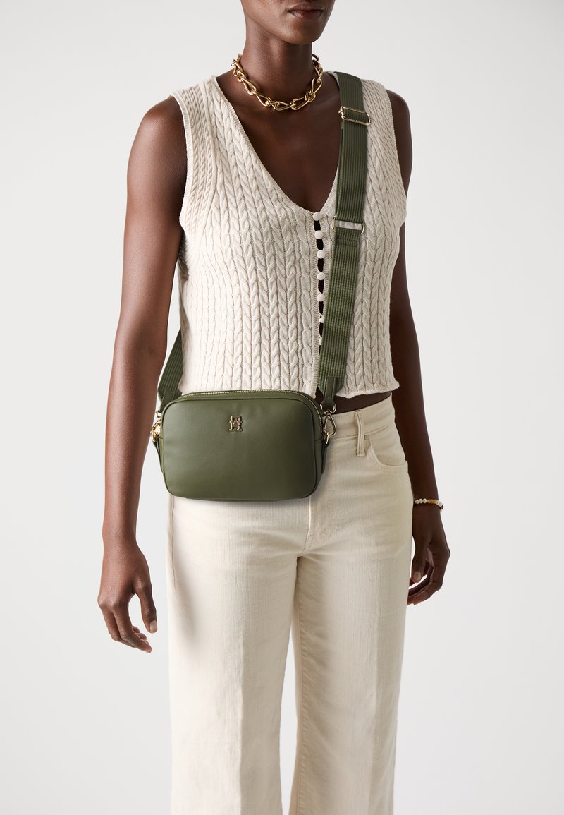 Olijfgroene crossbody tas met een rechthoekige vorm, gouden accenten en een gestructureerde schouderband. Draagt over een crèmekleurig gebreid bovenstuk en lichte broek.