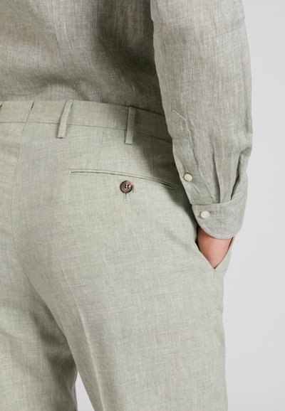 Hombre vistiendo pantalones de lino verde claro y camisa de manga larga a juego, con la mano apoyada en el bolsillo, mostrando el detalle del bolsillo trasero abotonado.