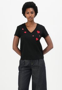Schwarzes Baumwoll-T-Shirt mit V-Ausschnitt, das auf der Vorderseite rot gestickte Herzchen in verschiedenen Größen präsentiert. Kurze Ärmel, taillierte Passform.