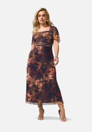 Kvinde i en brun og sort blomsterprint midi kjole med korte ærmer, firkantet halsudskæring og gyldne høje hæle sandaler, der står på en hvid baggrund.