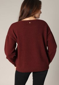Pull en tricot bordeaux avec une coupe décontractée, des manches larges, un ourlet côtelé et un petit accent doré au niveau de l'encolure arrière.
