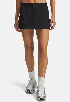 Zwarte, knie-lange rok met elastische tailleband, lichtgewicht materiaal, knoopsluiting aan de voorkant en zijzakdetail. Draagt zilveren sneakers.
