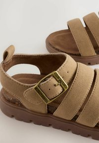 Braune Wildleder-Sandalen mit drei breiten Riemen, einer runden Zehenpartie und einer strukturierten braunen Sohle. Mit einer goldfarbenen Schnalle zur Anpassung.