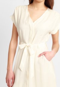 Vestido marfil con escote en V, mangas cortas, cintura ceñida con cinturón y bolsillos laterales. La tela parece suave y ligera.