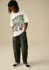 Weißes T-Shirt mit schwarzem und grünem Grafiktext, kombiniert mit dunkelgrauen Cargo-Hosen mit Seitentaschen. Schwarze Sneakers mit weißen Akzenten.