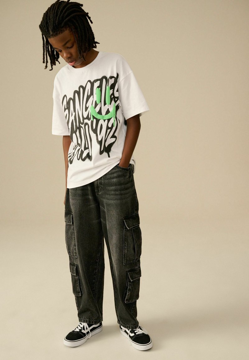 Weißes T-Shirt mit schwarzem und grünem Grafiktext, kombiniert mit dunkelgrauen Cargo-Hosen mit Seitentaschen. Schwarze Sneakers mit weißen Akzenten.