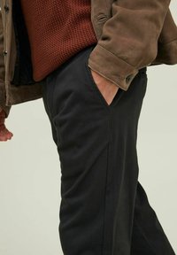 Chaqueta de ante marrón sobre un suéter texturizado color óxido y pantalones negros. Detalle del bolsillo visible en los pantalones con un ligero brillo.